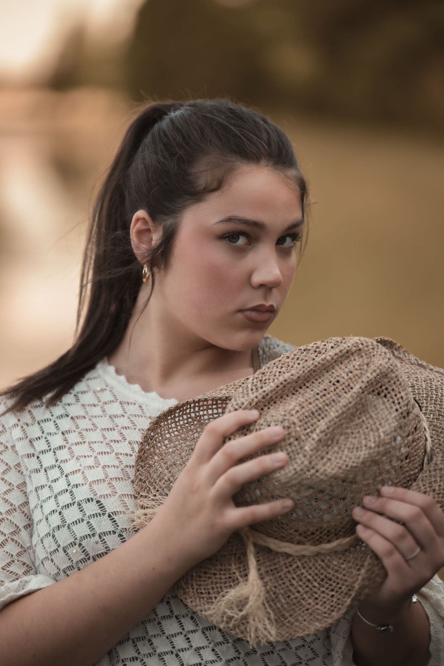 Young woman with dark ponytail holding burlap fabric outdoors, wearing white lace top in golden hour light.