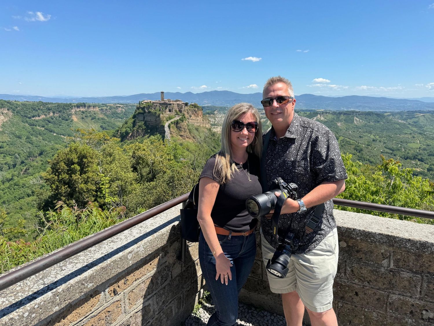 People enjoying scenic mountain views from a stone overlook on a sunny day with green landscapes in the background.