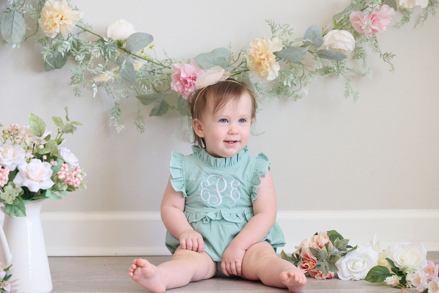 Baby in mint green romper sits on floor surrounded by delicate floral decorations and soft pink accents.