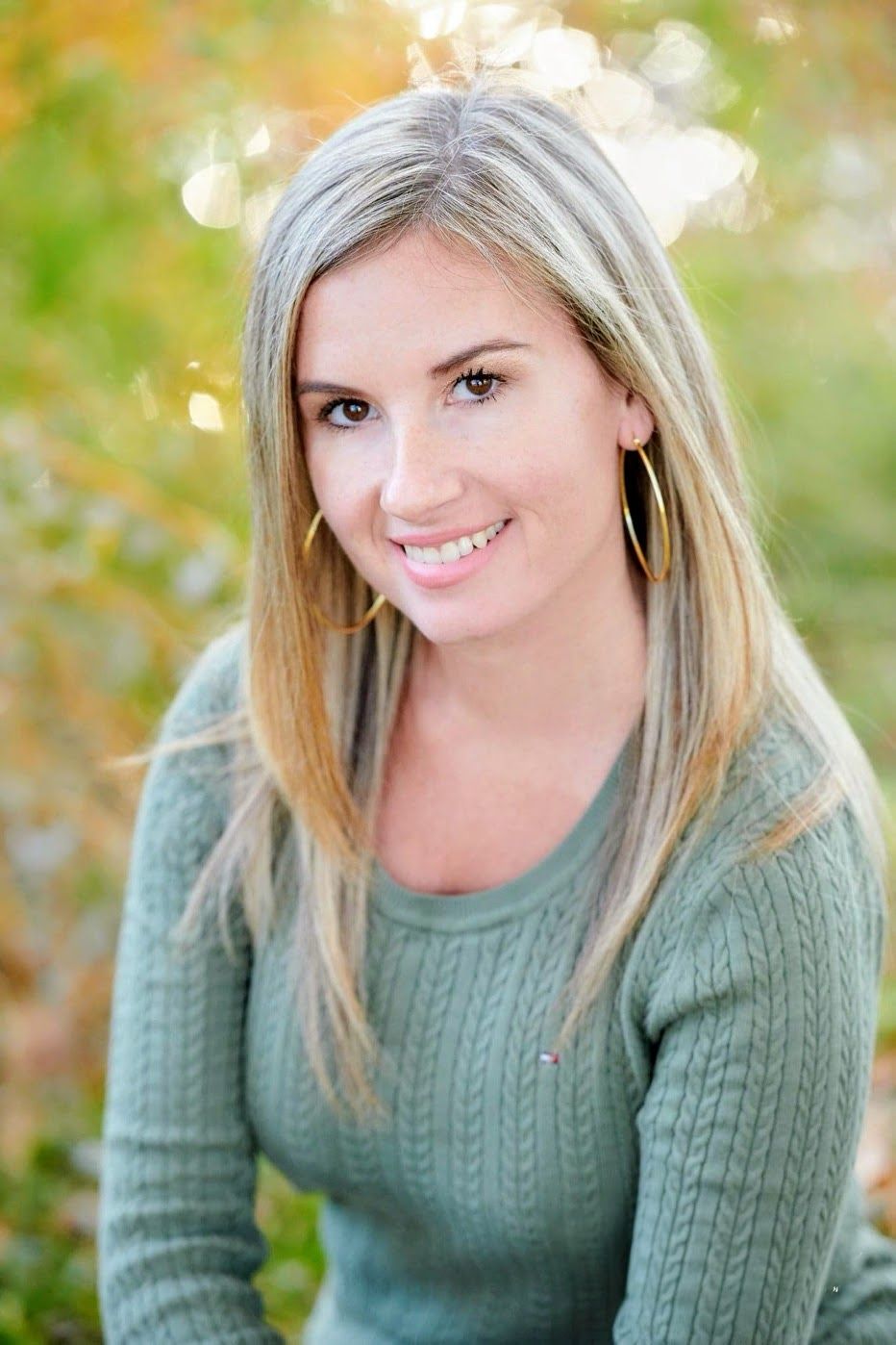 Portrait photo with soft bokeh background showing someone in a sage green cable knit sweater and gold hoop earrings.