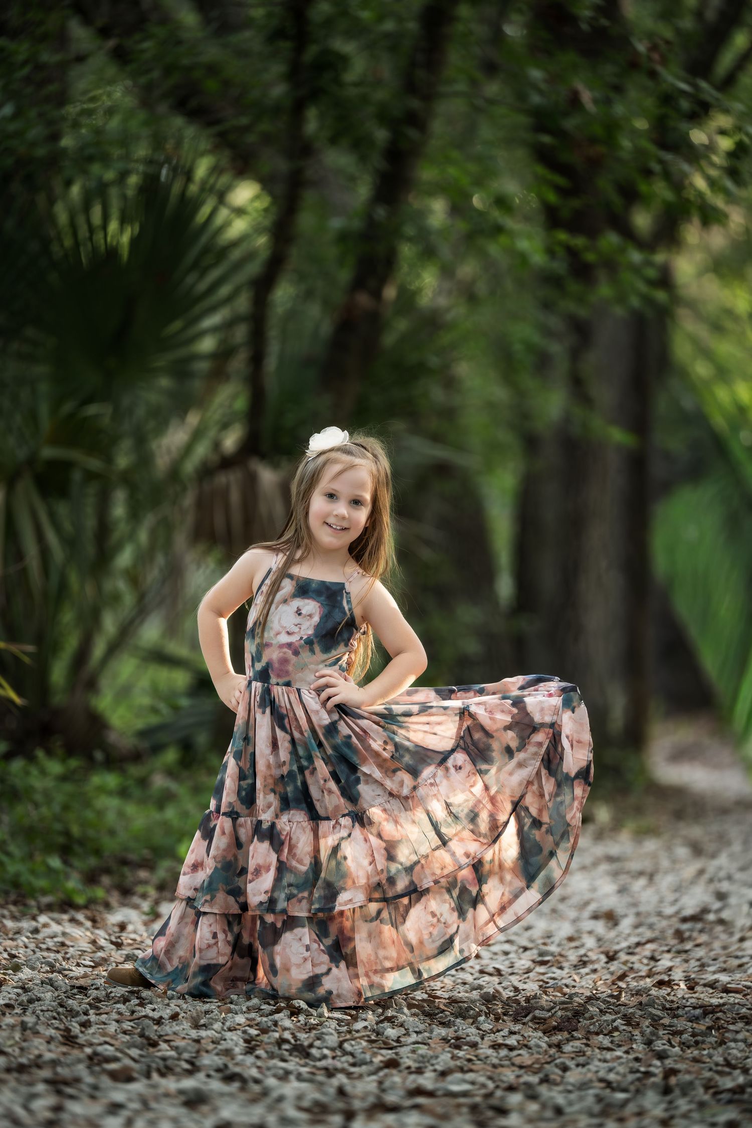 Young girl in floral maxi dress twirling on a gravel path surrounded by lush green trees.