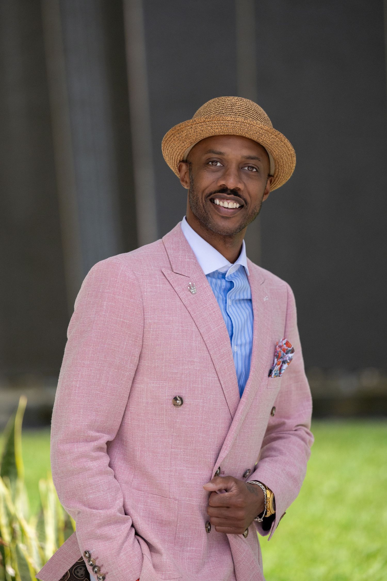 Dapper man wearing pink suit, straw fedora, and blue striped tie holding a clutch outdoors.