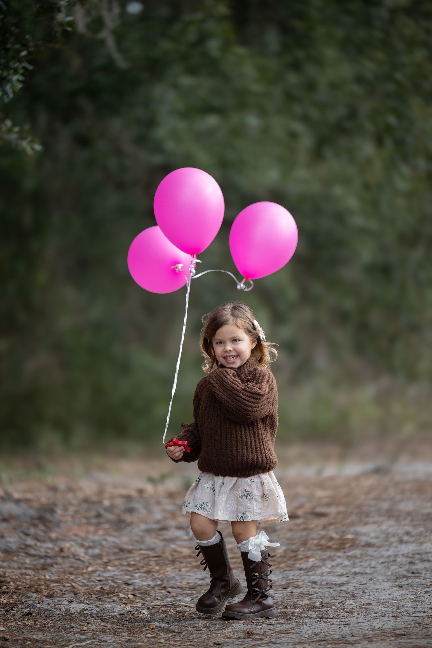 Child in brown knit hat and dress playing with bright pink balloons on a forest path.