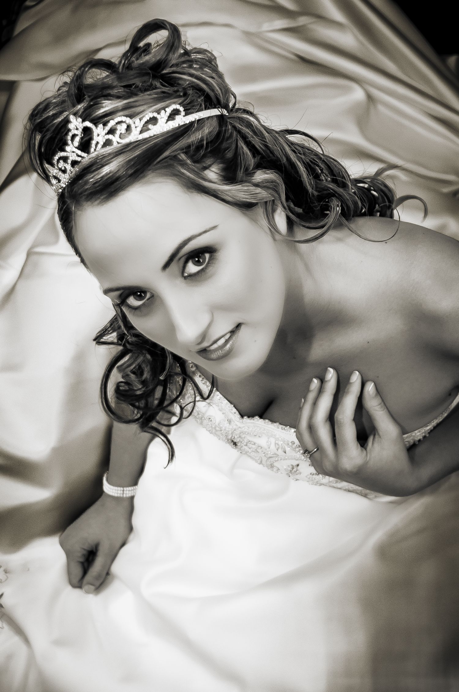 Black and white portrait of a bride wearing a delicate tiara and white dress laying on satin fabric.