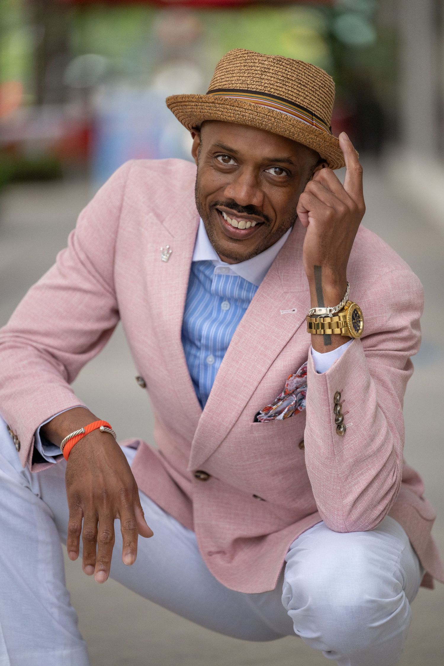 Stylish man in pink blazer, straw hat, and gold watch smiling confidently outdoors.