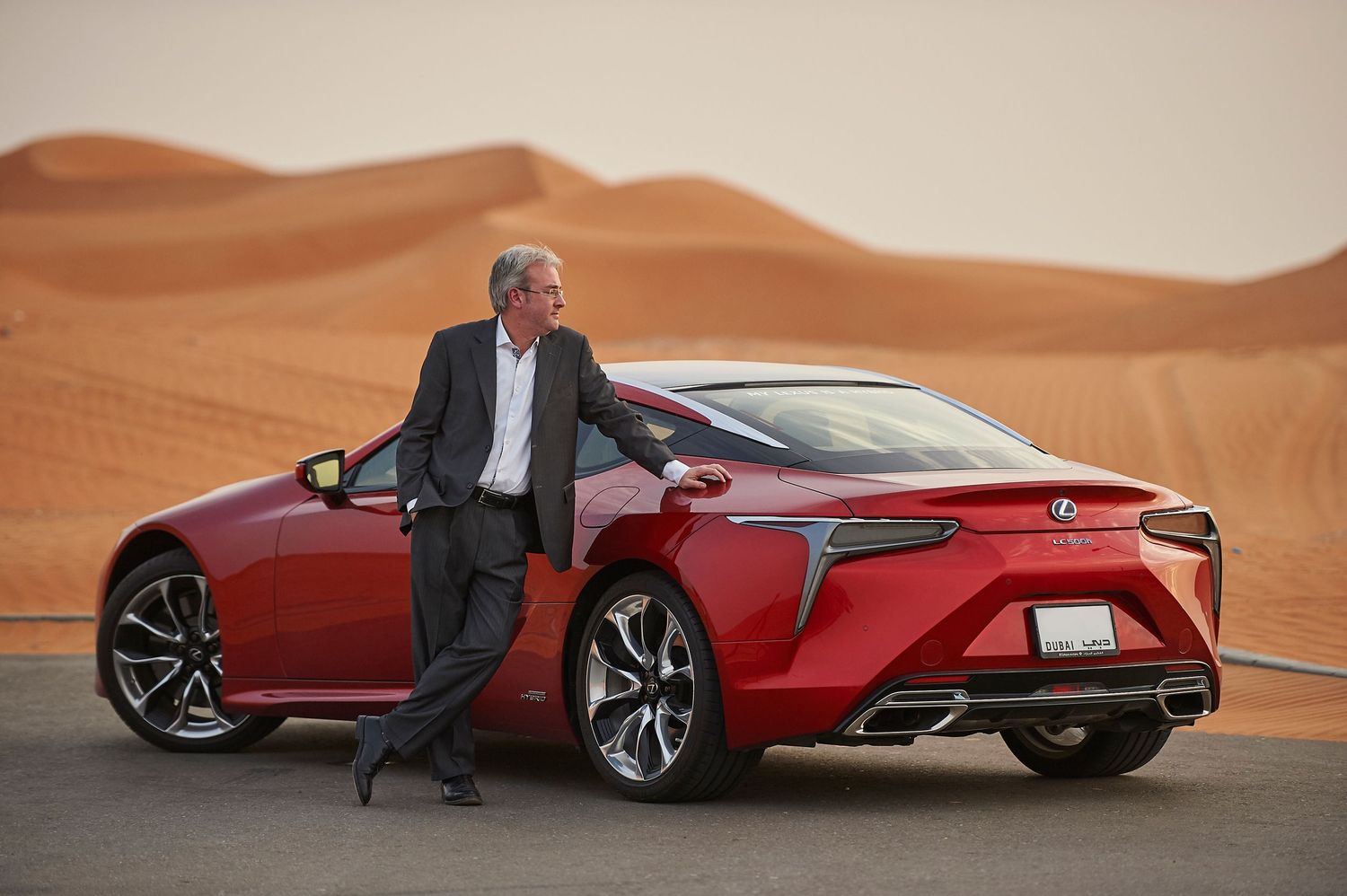 Red Lexus LC sports car parked in desert sand dunes at sunset.