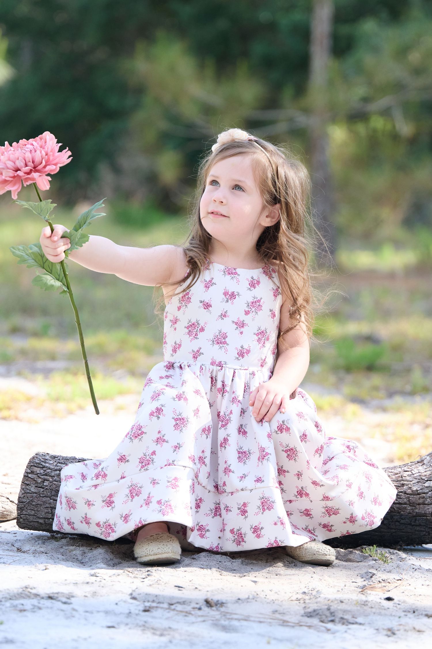 Child in floral print dress sits on log while reaching toward pink flower in outdoor garden setting.