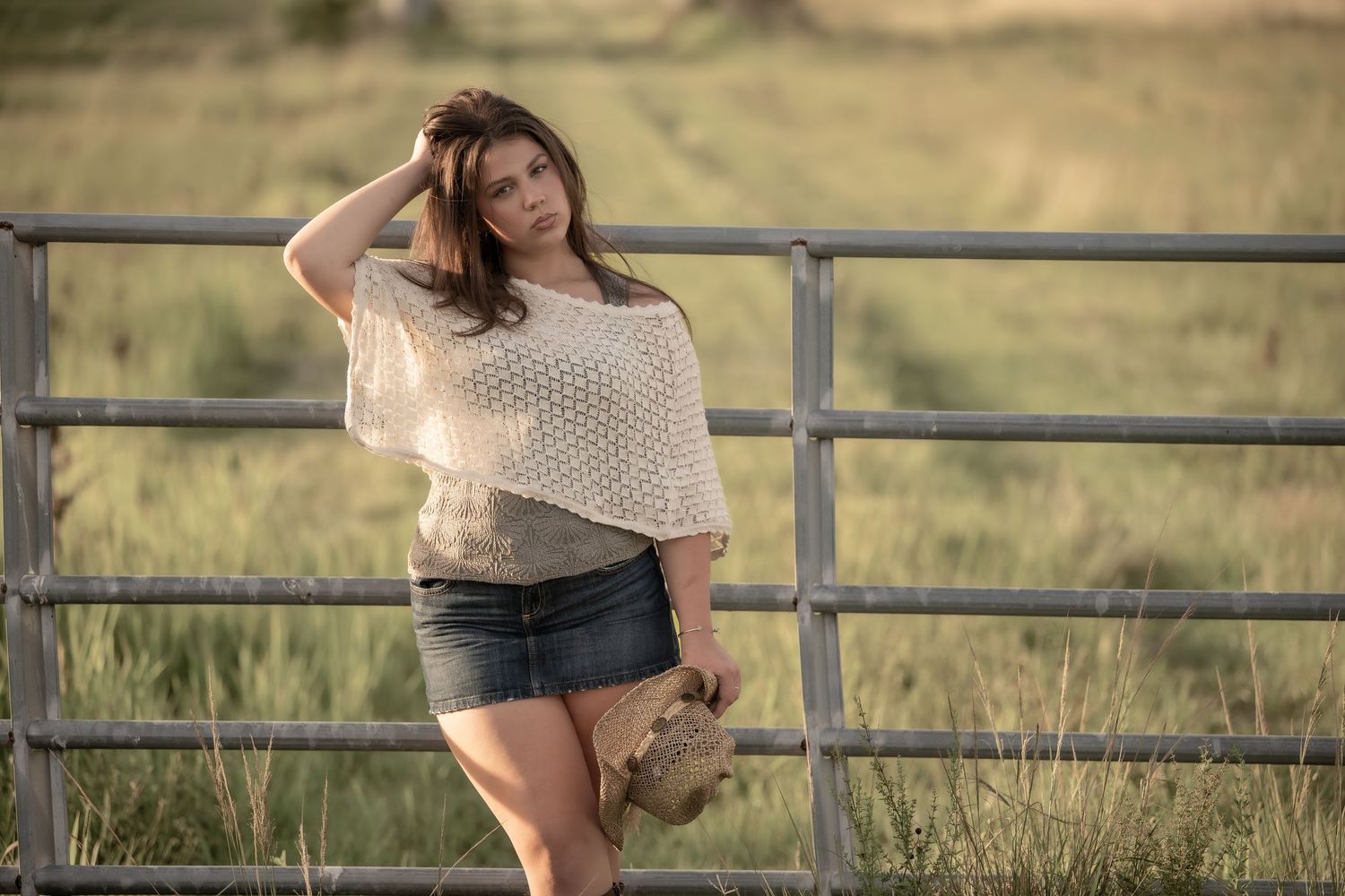 Young woman in white knit top and denim skirt poses by a rustic metal farm gate in a golden field.