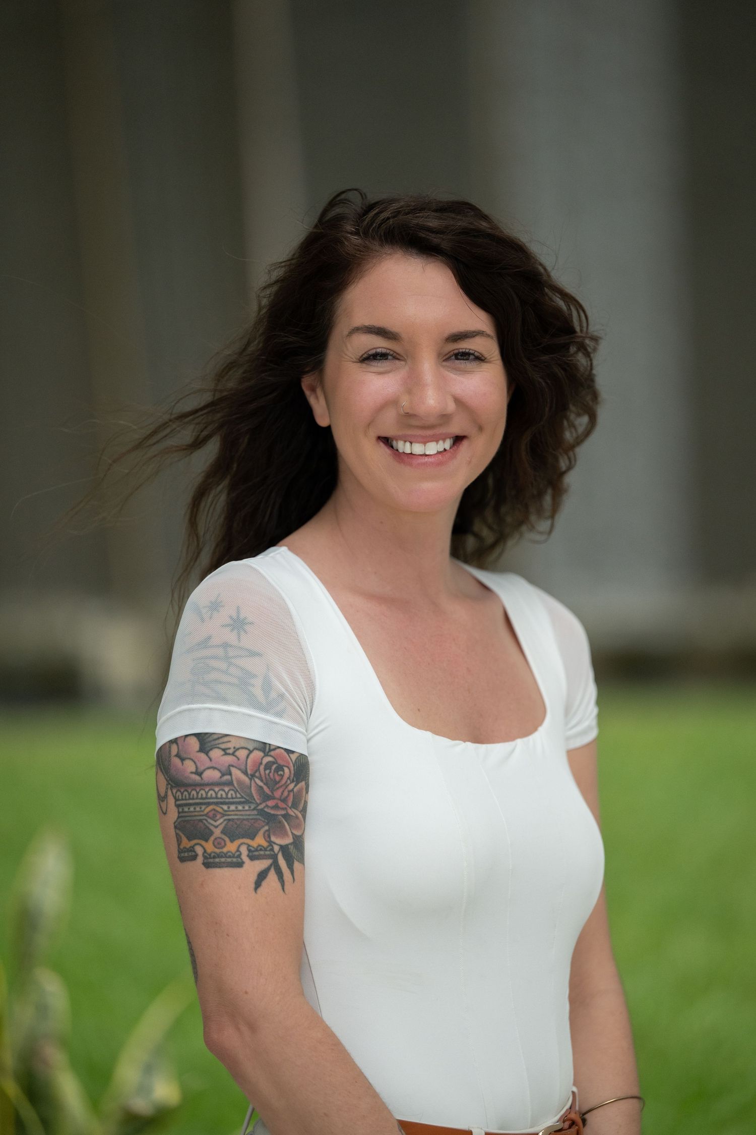 Smiling woman with dark curly hair and floral arm tattoo wearing white top outdoors.