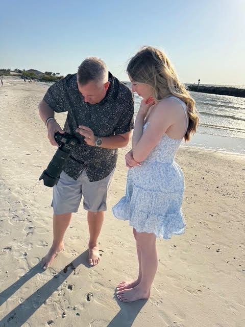 Two people bend down to collect seashells together on a sunny beach by the ocean.