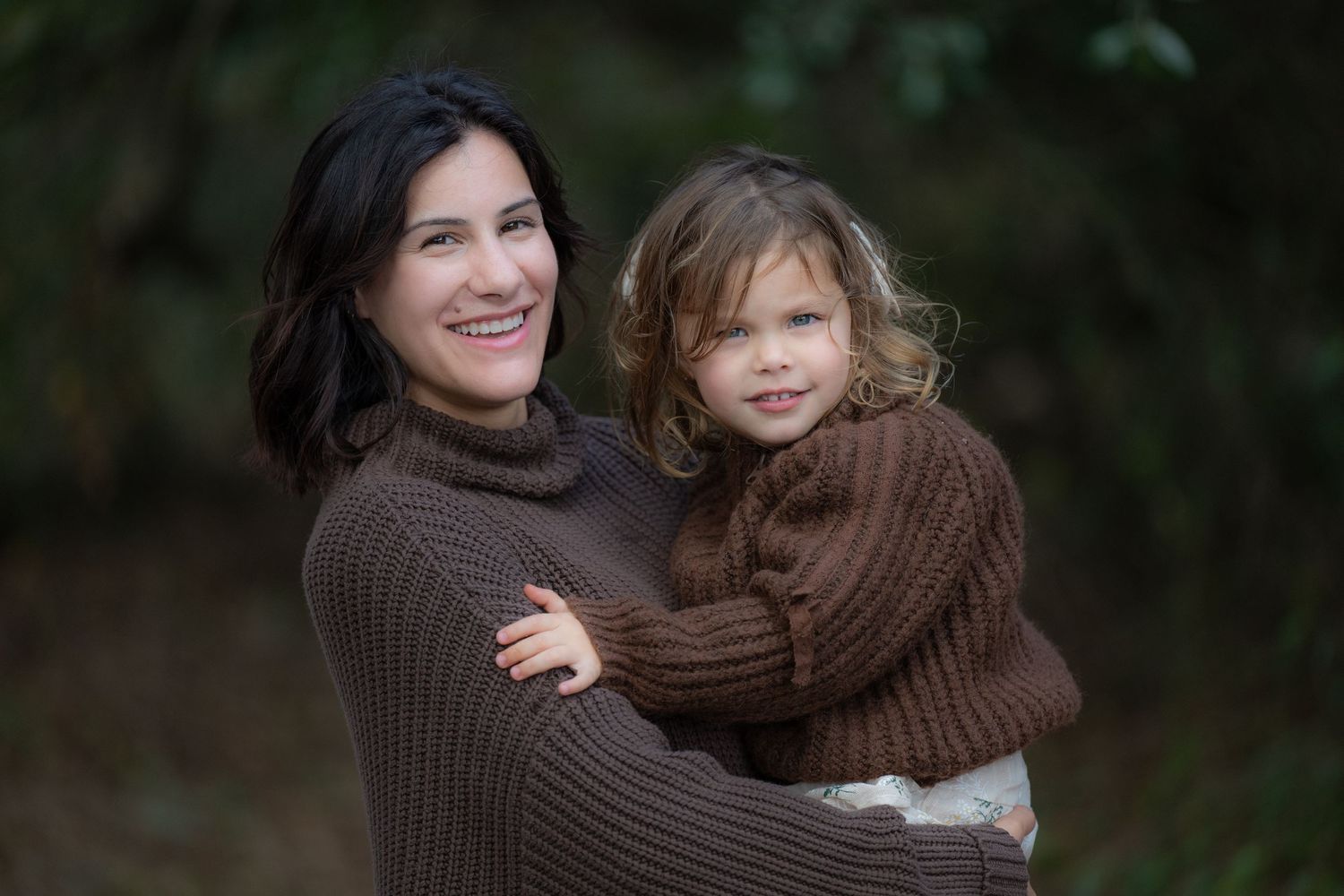 A parent and child wearing matching brown sweaters embrace while smiling outdoors against a dark background.