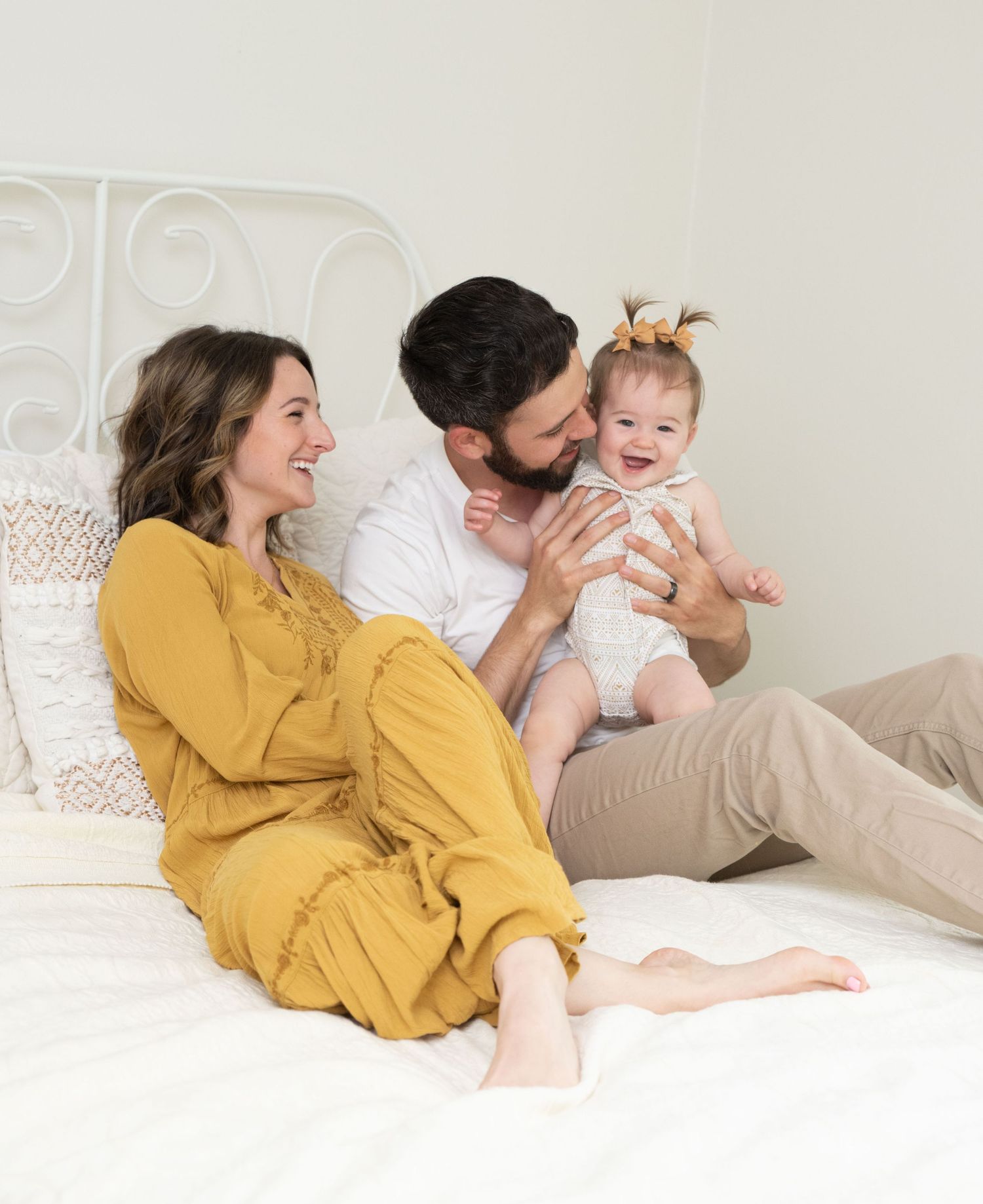 Family lounging together on white bedding wearing yellow and neutral colors in a bright, airy bedroom setting.