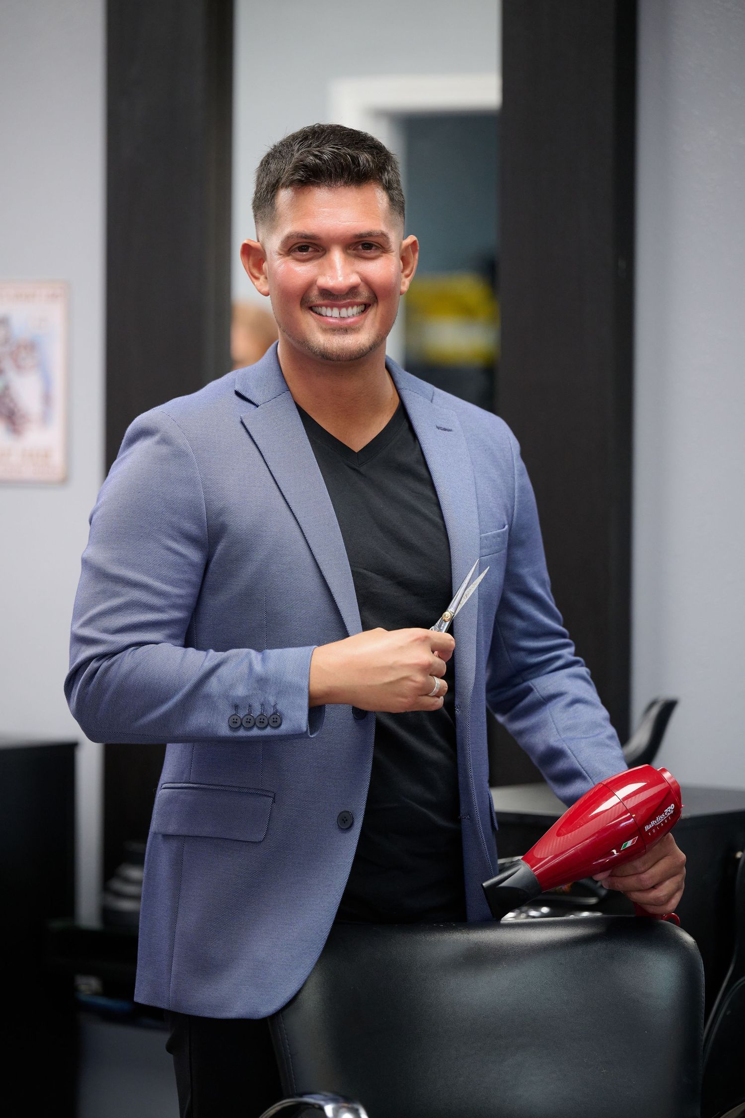 A person in a blue blazer and black shirt smiles while holding a red coffee maker in an office setting.