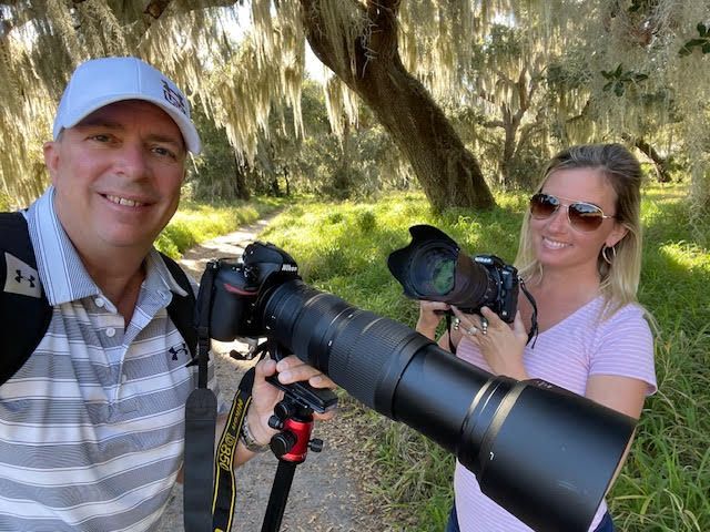 Two photographers with professional cameras and telephoto lenses stand together in a sunny outdoor setting.