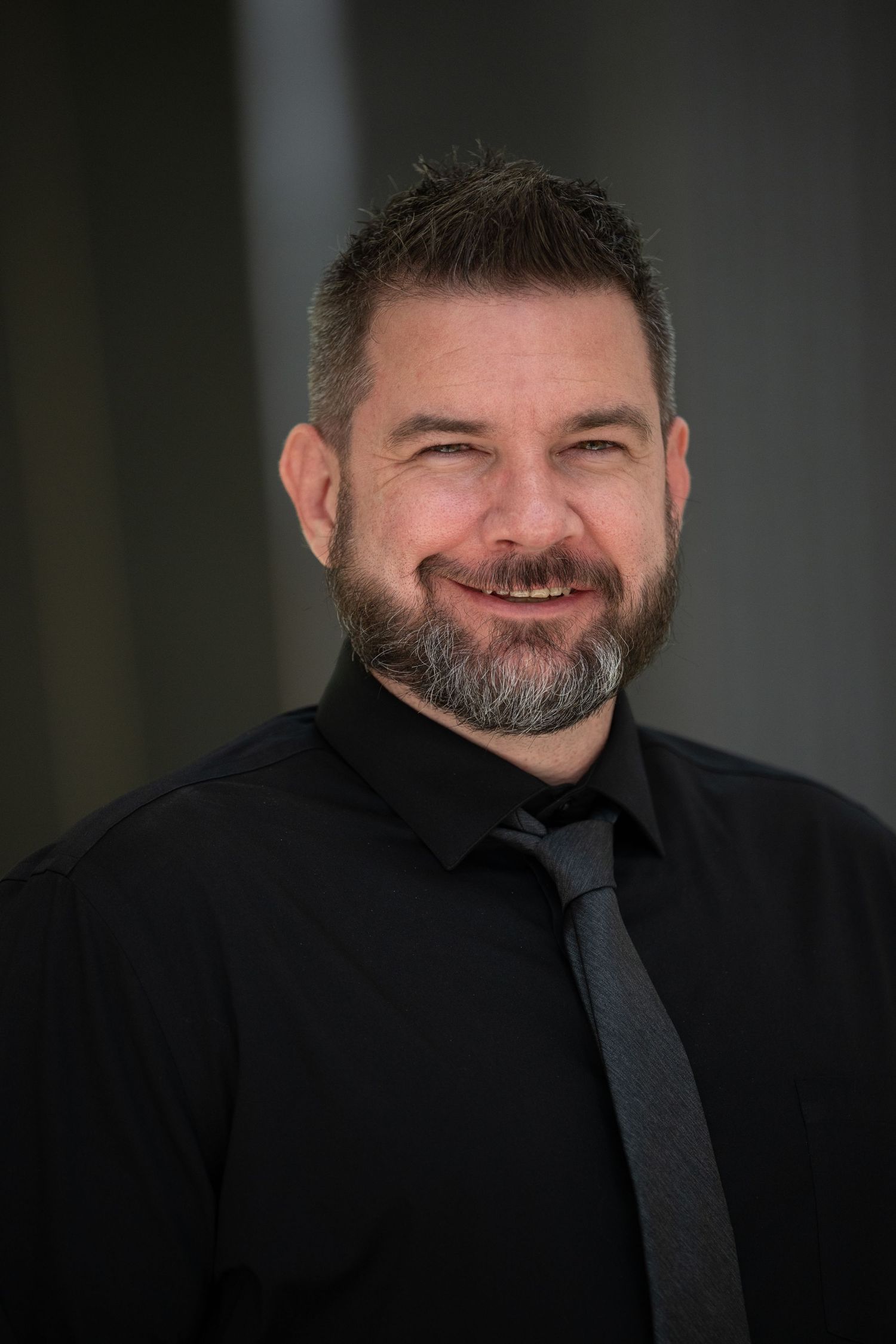 Bearded man in black shirt and tie smiling warmly in a professional headshot.