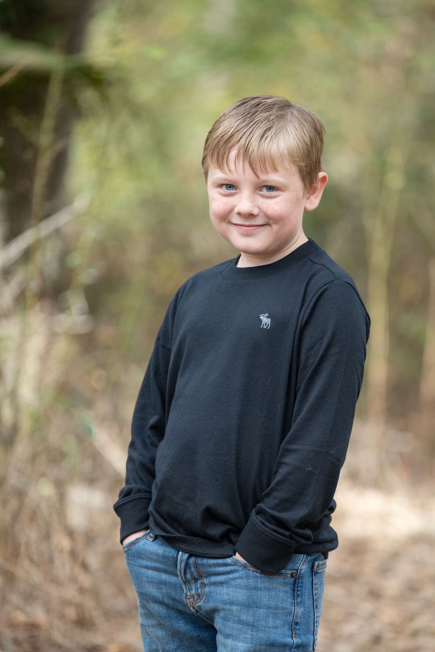A young person in a black sweater and jeans poses outdoors against a blurred natural background.