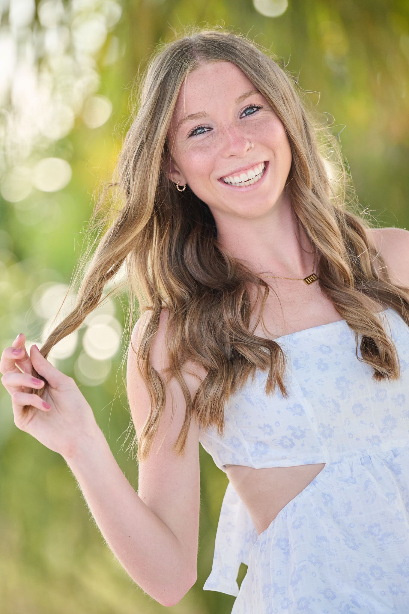 A person in a white lace top poses playfully against a soft green bokeh background outdoors.