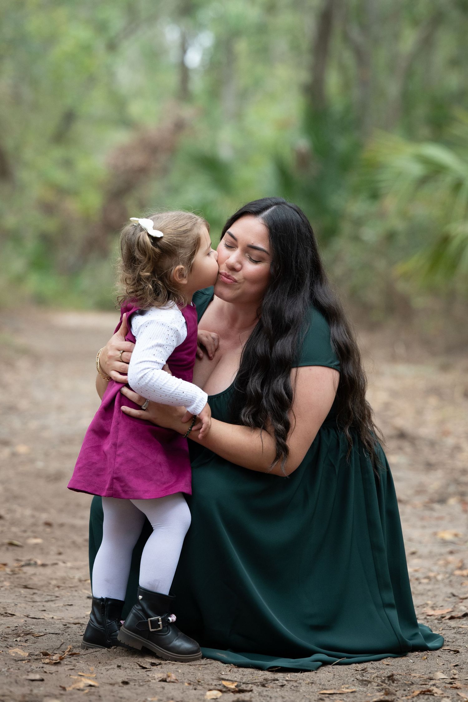 A mother and daughter share tender moments together outdoors in a forest setting wearing green and purple dresses.