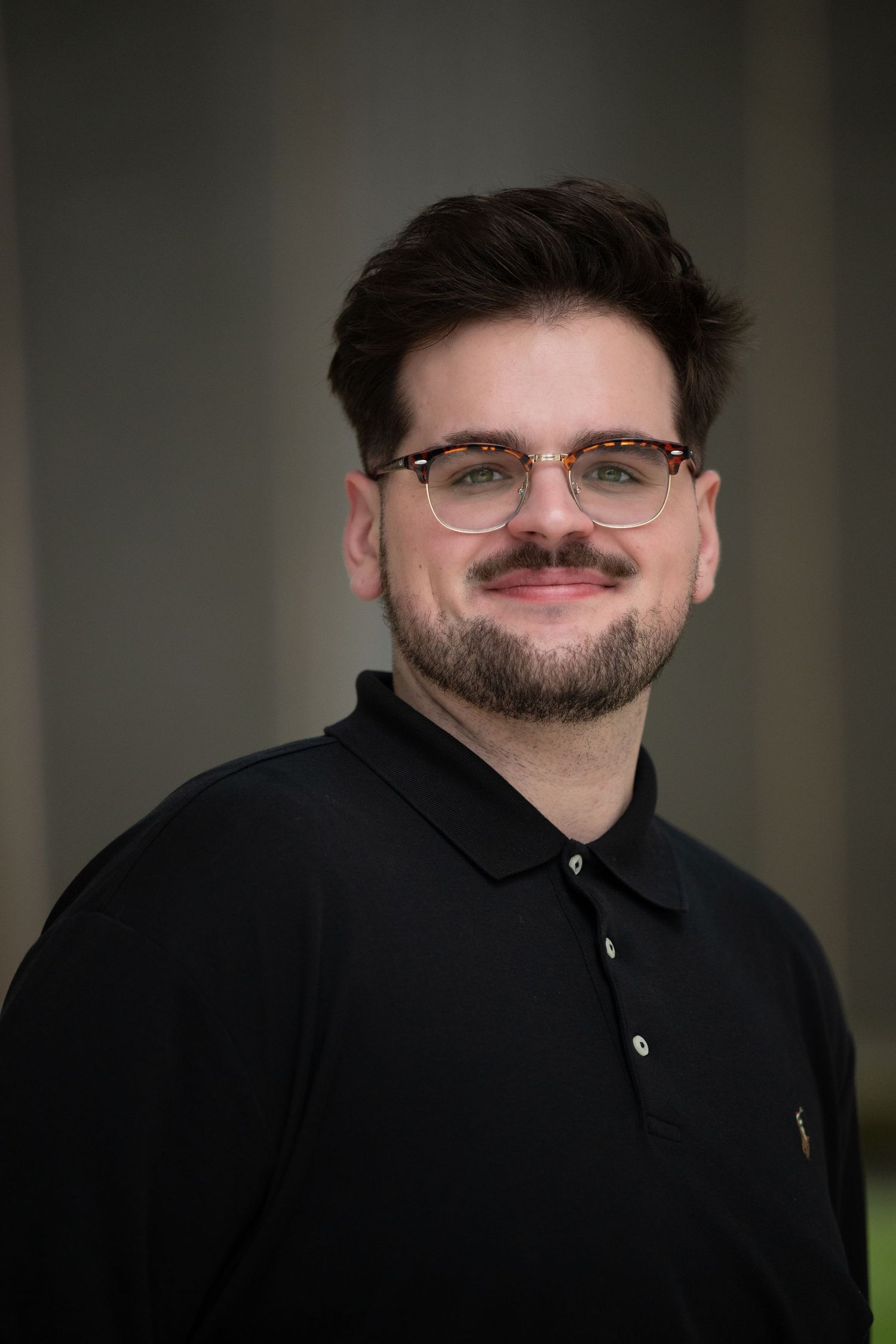 Young bearded man with glasses wearing a black polo shirt in a professional headshot.