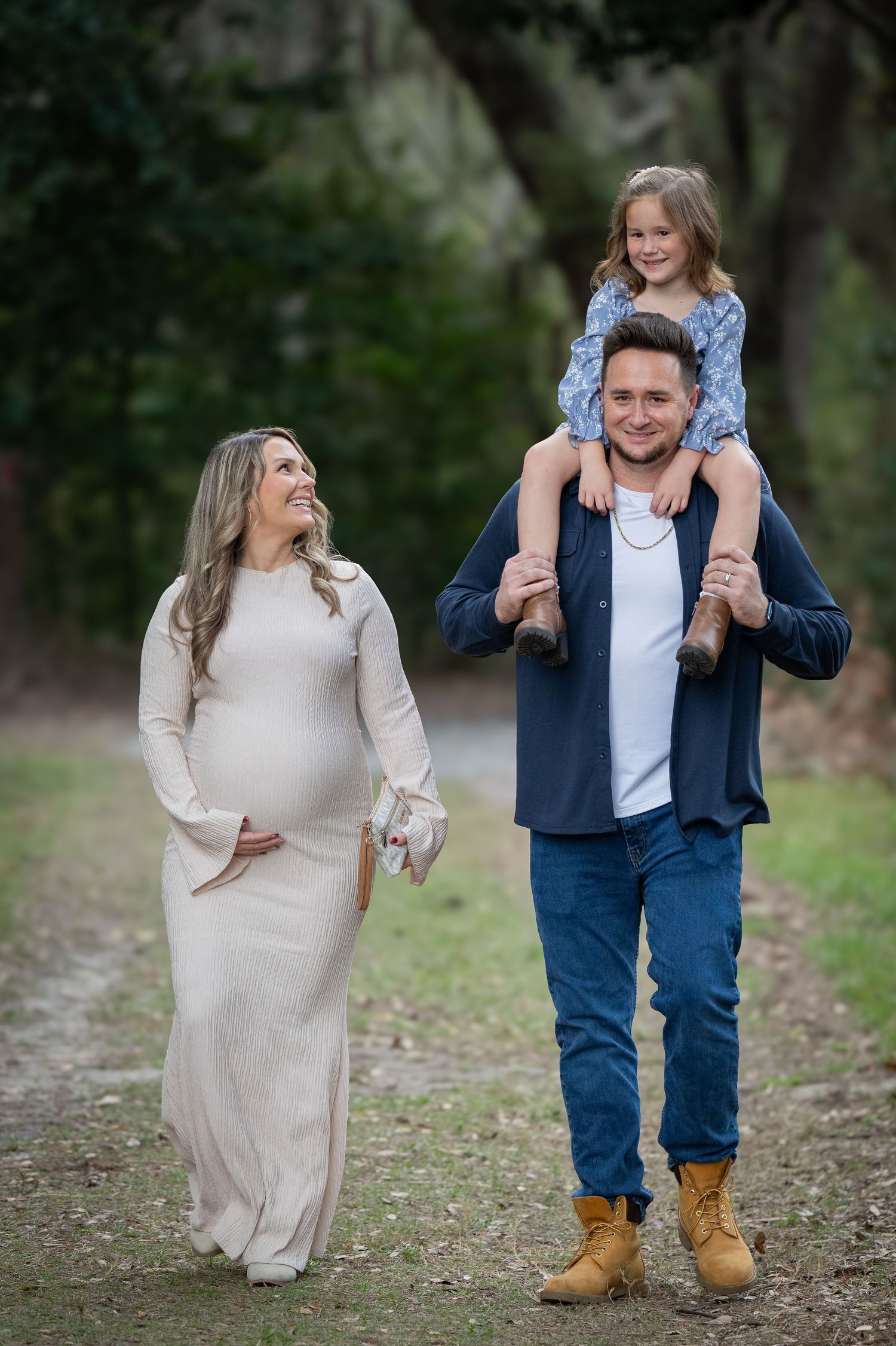 A couple walks together on a path through a wooded area while a child rides on shoulders in a playful moment.