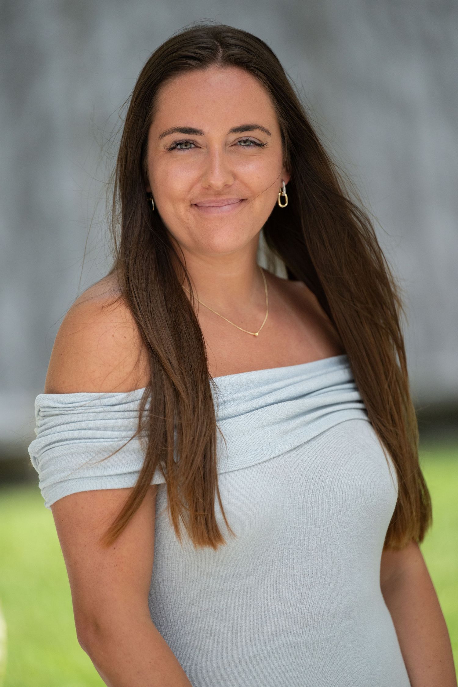 Brunette woman in light blue off-shoulder top smiling softly outdoors.