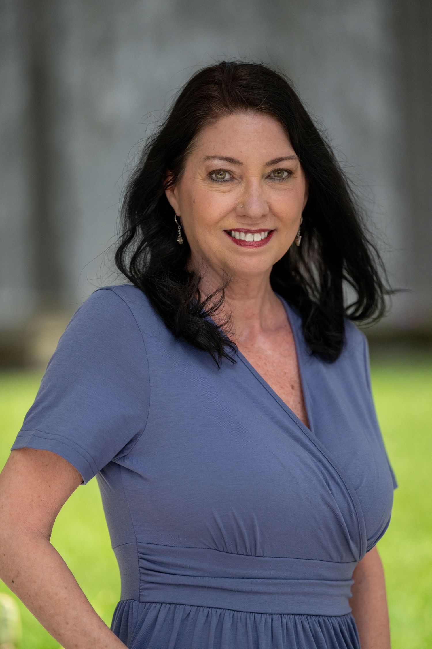 Smiling woman with dark hair wearing a periwinkle wrap dress posing outdoors.