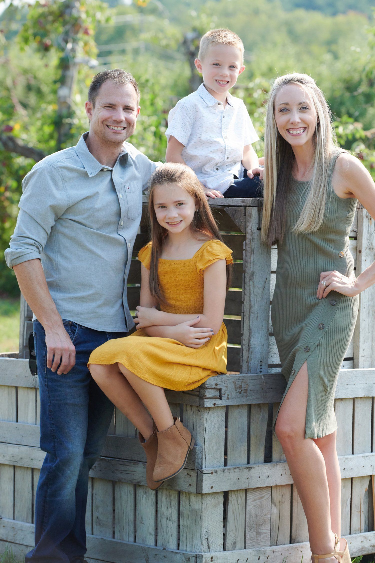 A family poses casually along a wooden fence with trees and greenery in the background.