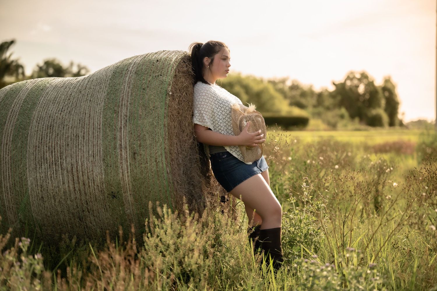 Young woman in denim shorts and boots leaning against a round hay bale in a sunlit field.