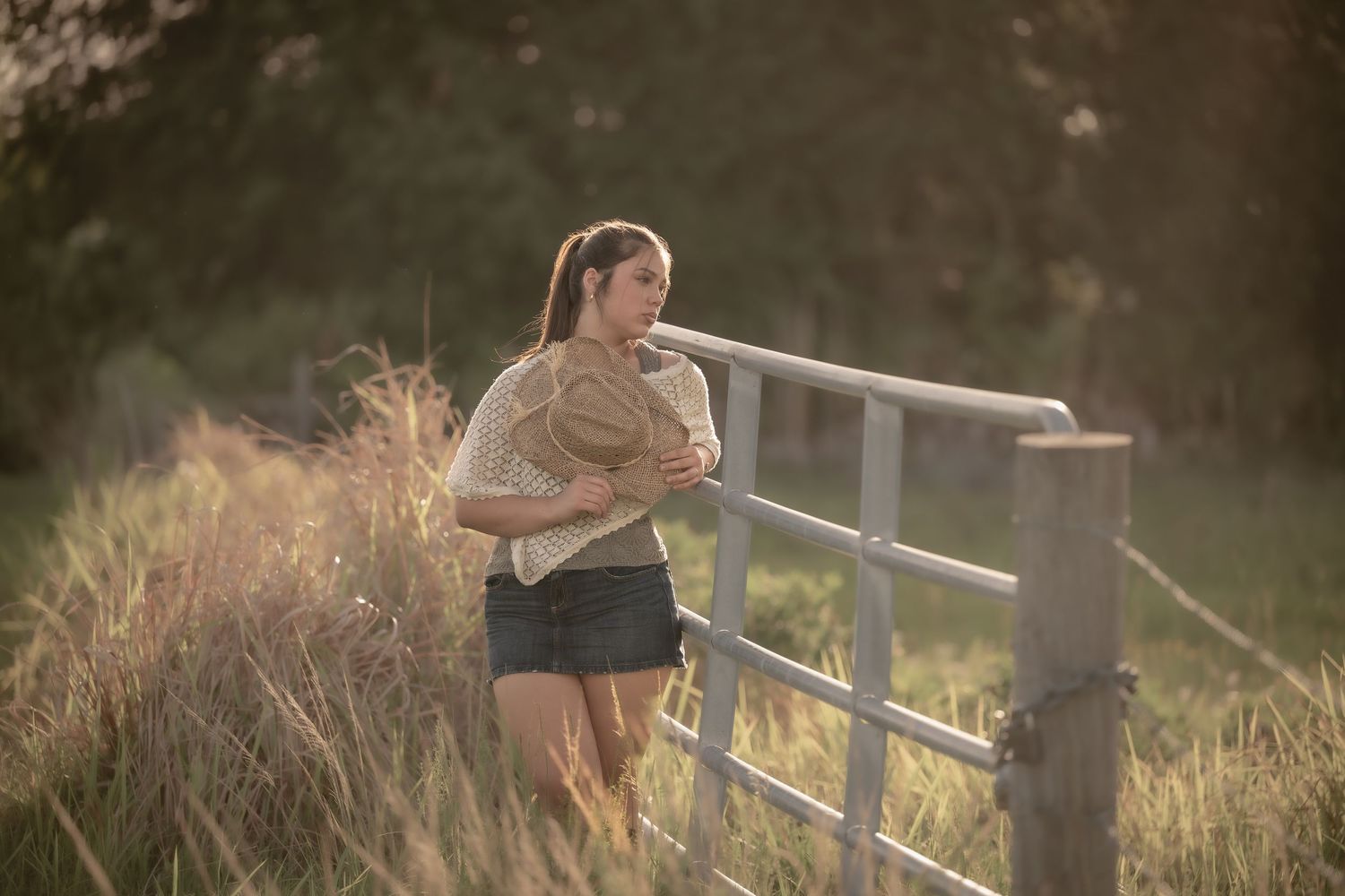 Young woman in casual outfit leaning on a wooden fence in a golden sunlit field at sunset.