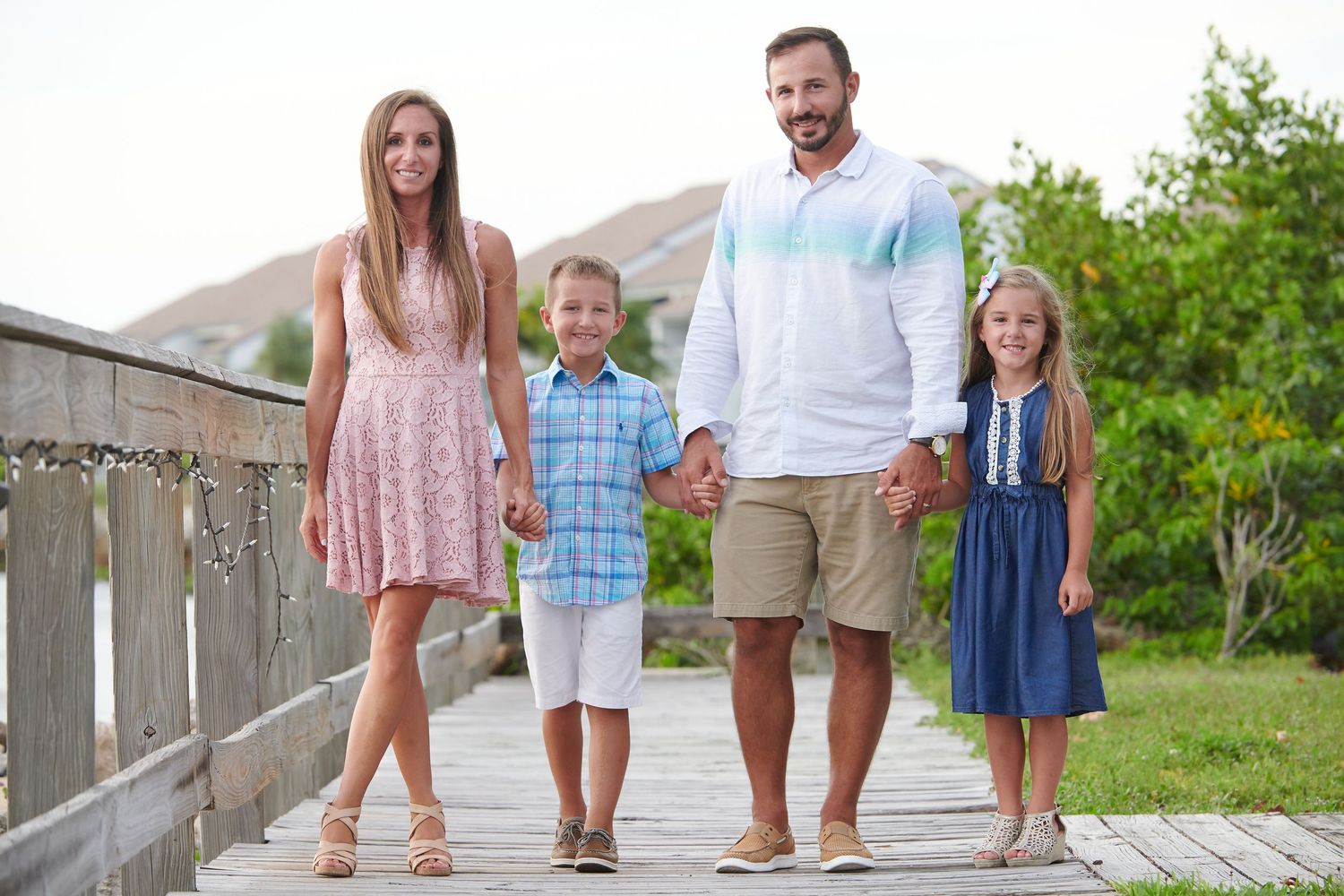 Family wearing coordinated blue and white outfits poses together on wooden boardwalk at beach during sunset.