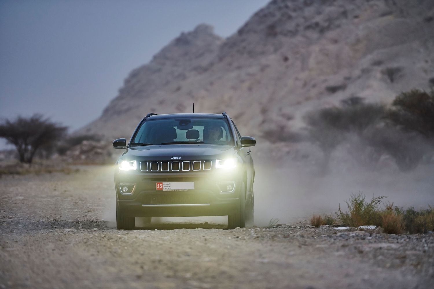 Front view of Jeep Compass driving through dusty desert terrain with headlights on.