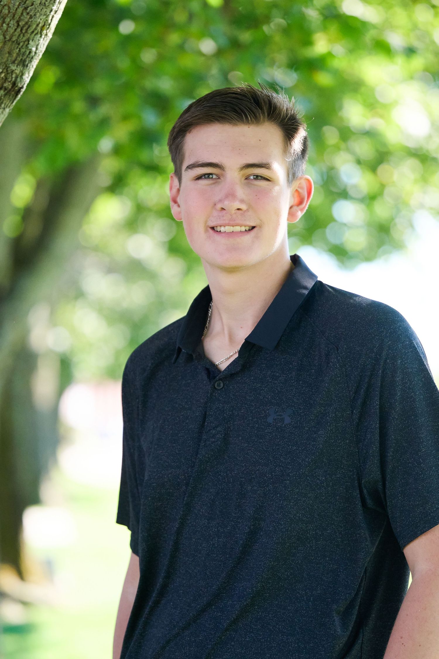 A person wearing a black polo shirt poses outdoors with trees and greenery in the background.