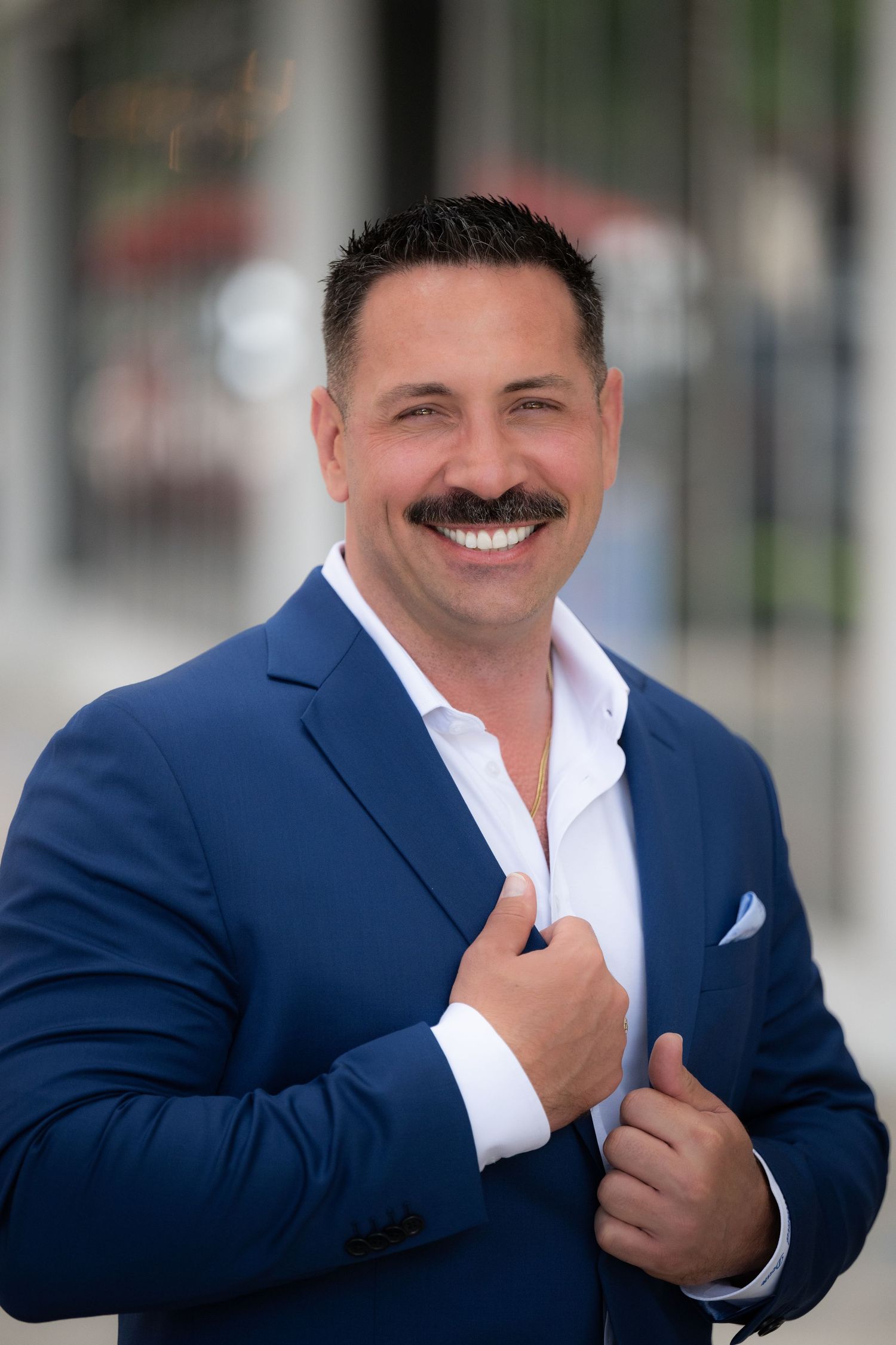 Smiling man with mustache in blue blazer and white shirt posing outdoors in a professional portrait photo.