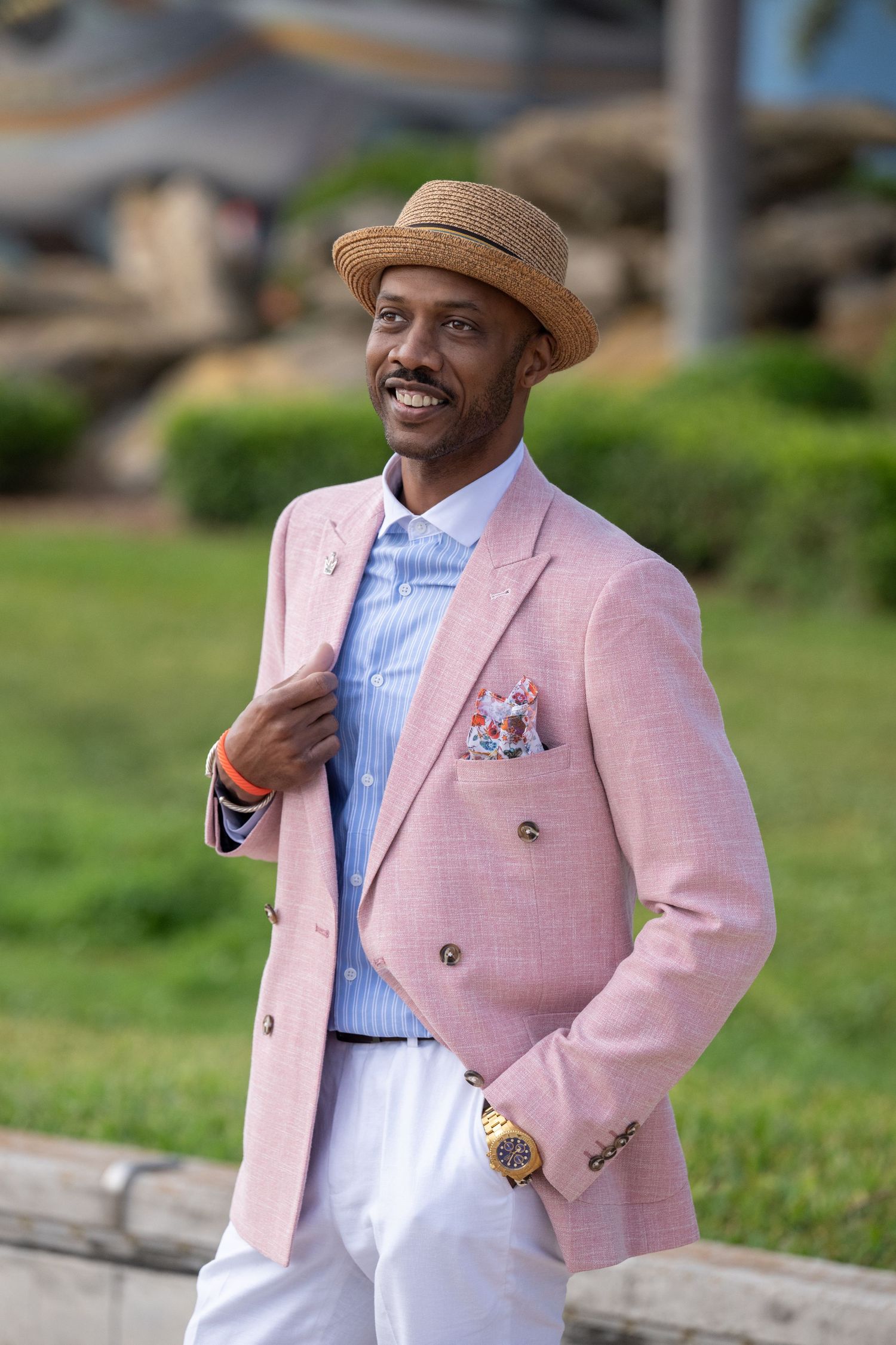 Stylish man in pink double-breasted blazer, straw hat, and floral pocket square smiling outdoors.