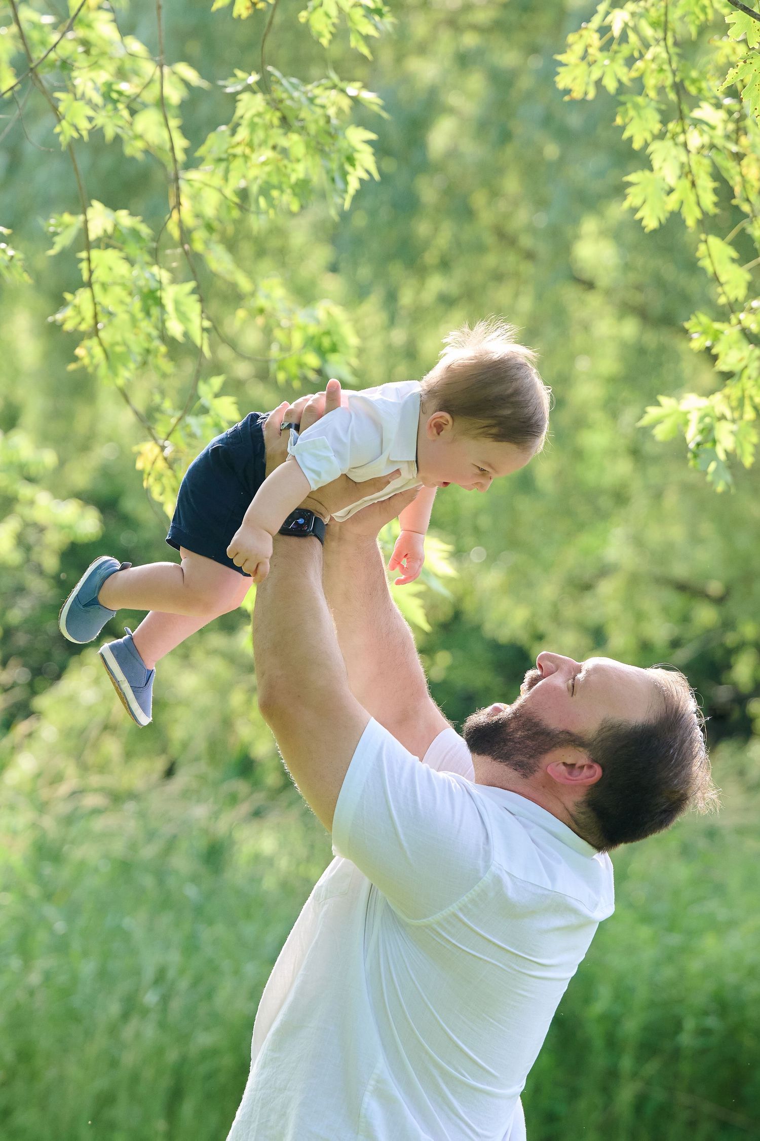 Parent playfully lifts small child in air against backdrop of sunlit green foliage.