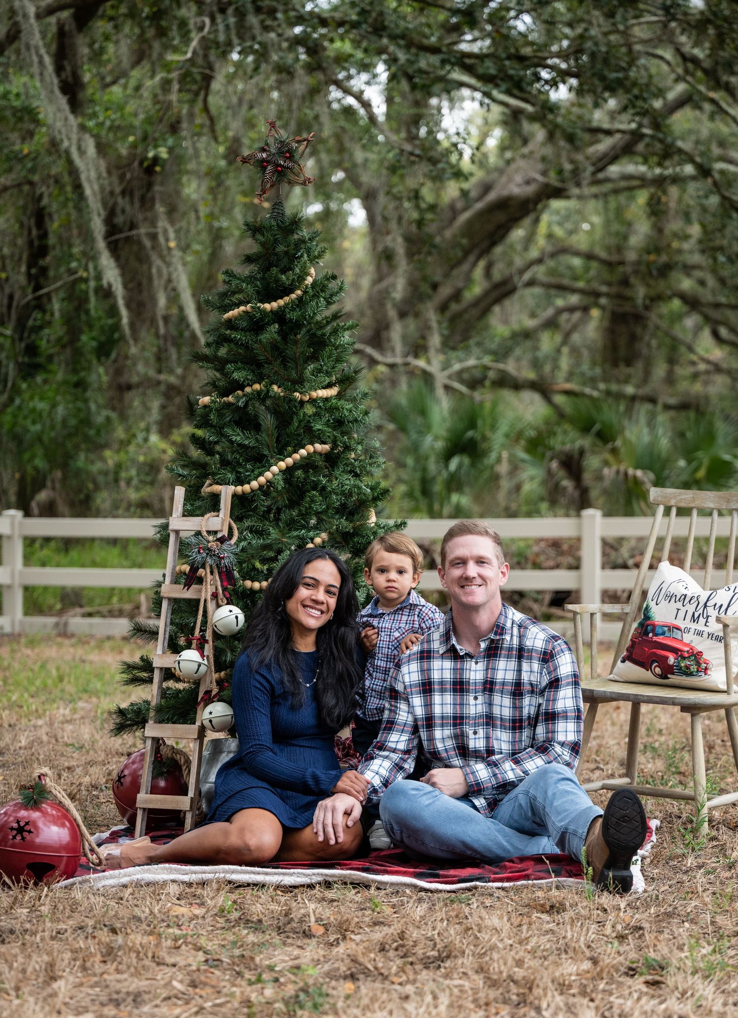 A family sits on a blanket by a decorated Christmas tree outdoors with rustic wooden fence and forest backdrop.
