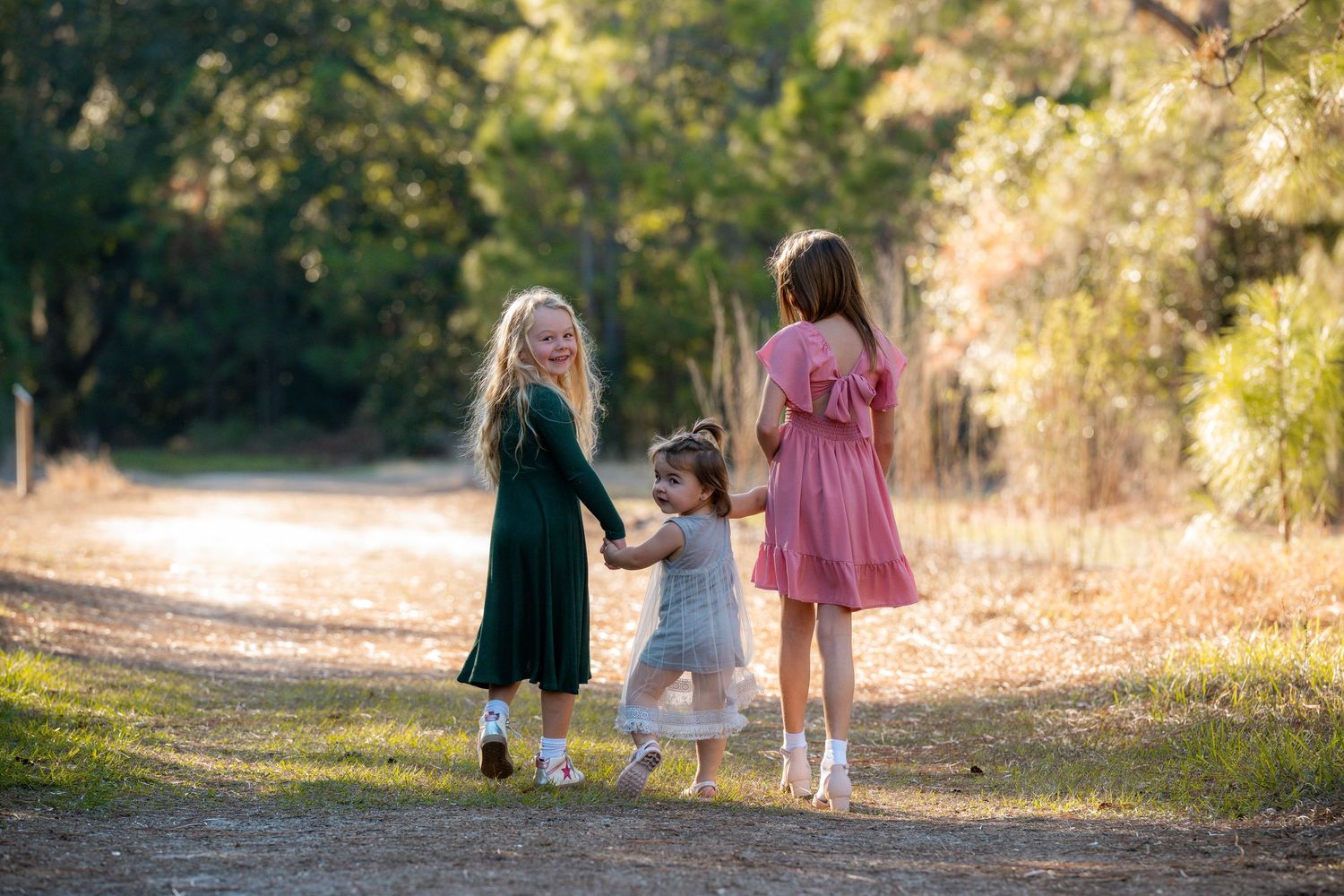 Three people walking together on a sunlit nature path wearing dresses in green, gray and pink colors.