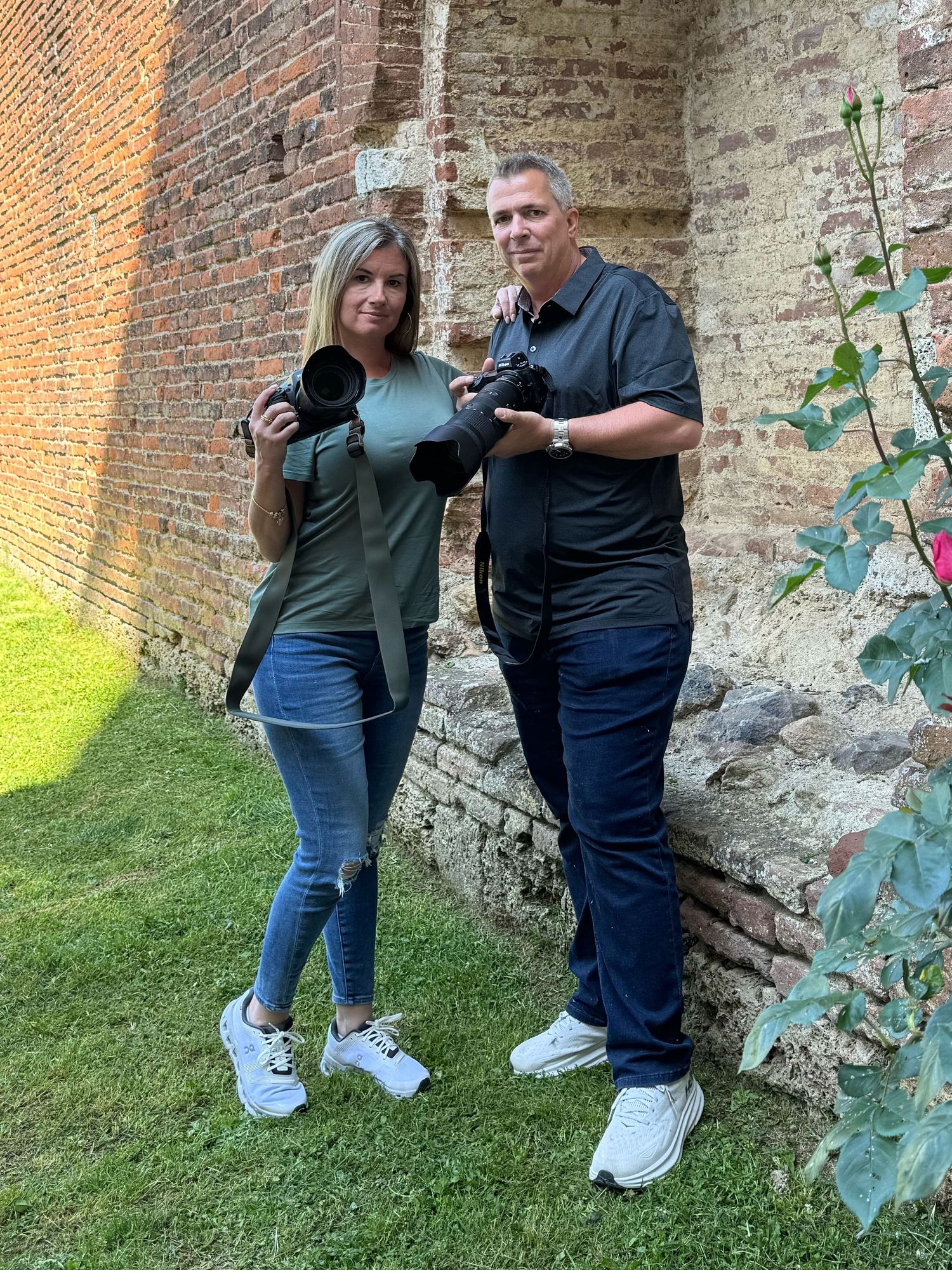 Two photographers with DSLR cameras pose together against a rustic brick wall outdoors.