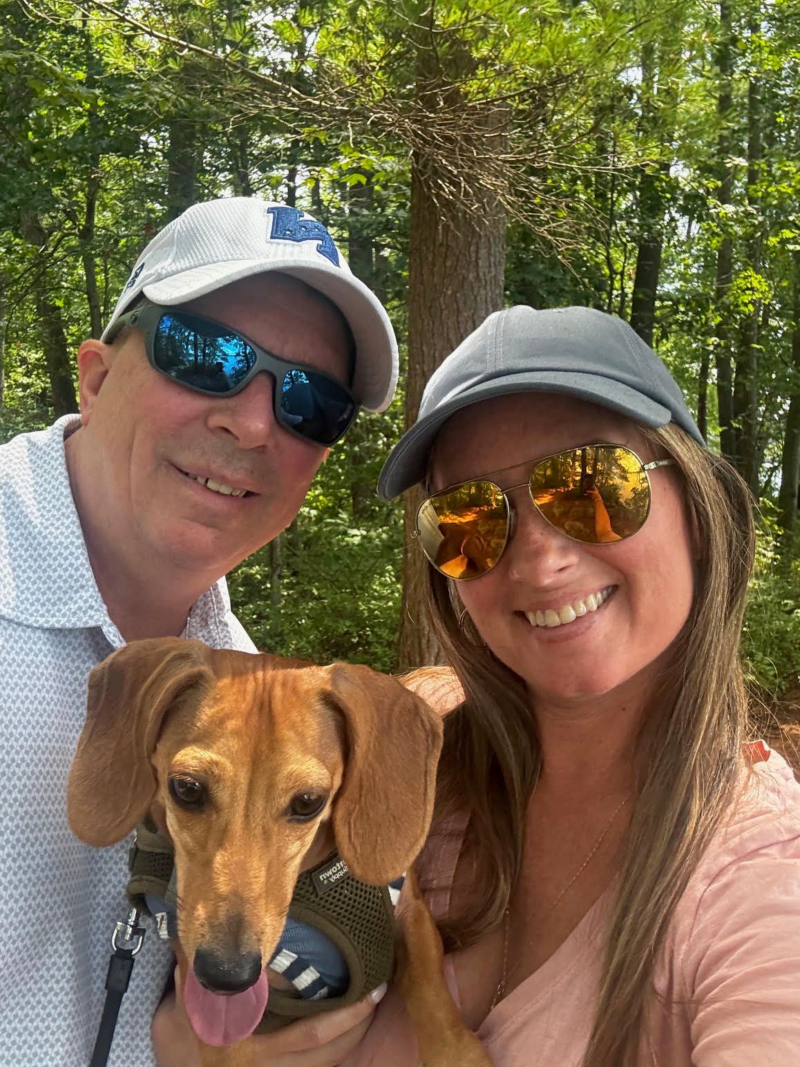 A couple poses outdoors with their brown dachshund dog in a series of selfies taken in a wooded setting.