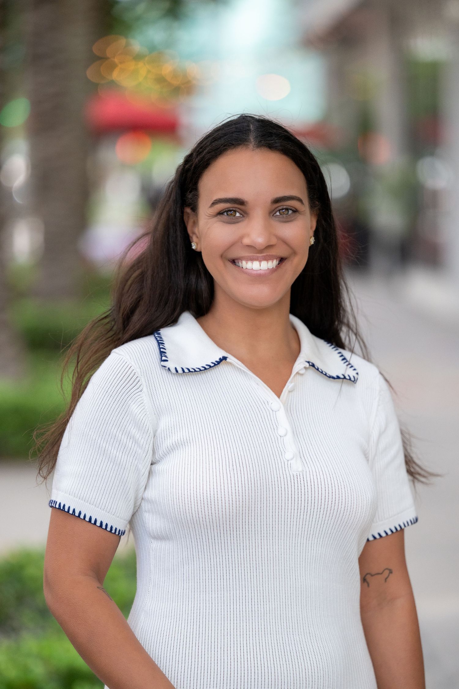 Smiling young woman with long dark hair wearing a white ribbed polo shirt with blue trim, standing outdoors.