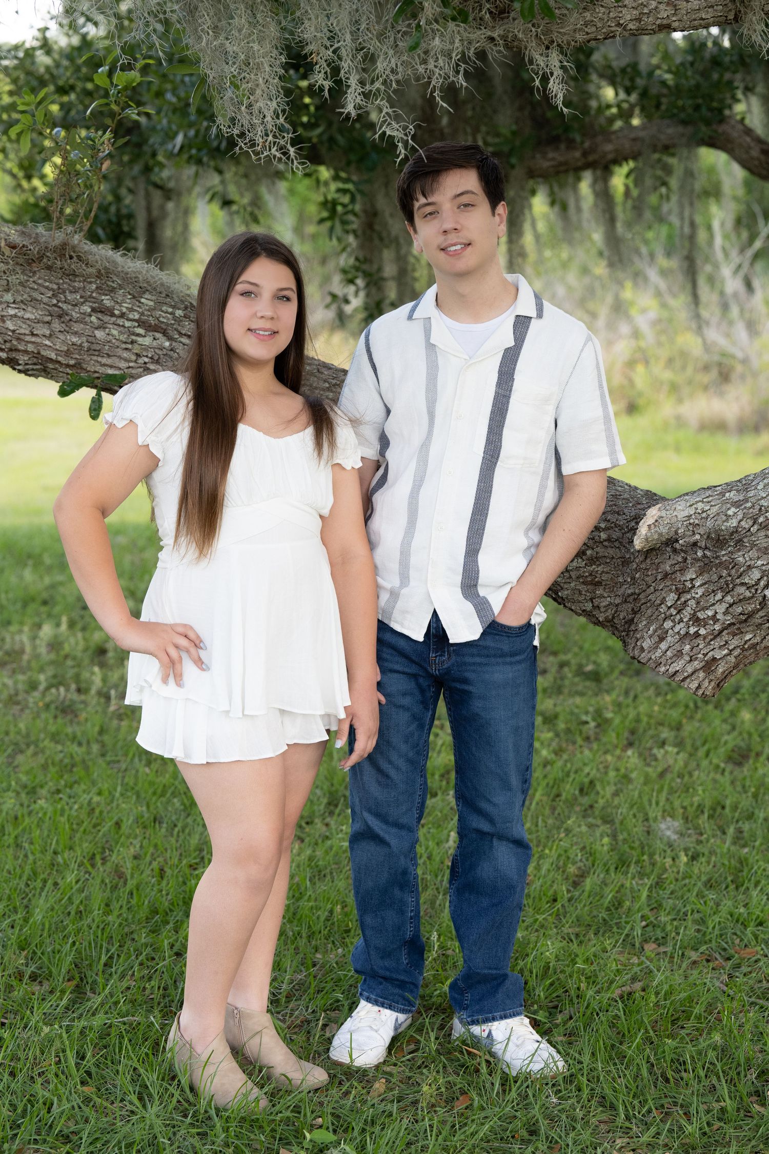 A couple in white and denim outfits poses playfully in a grassy outdoor setting with trees in the background.