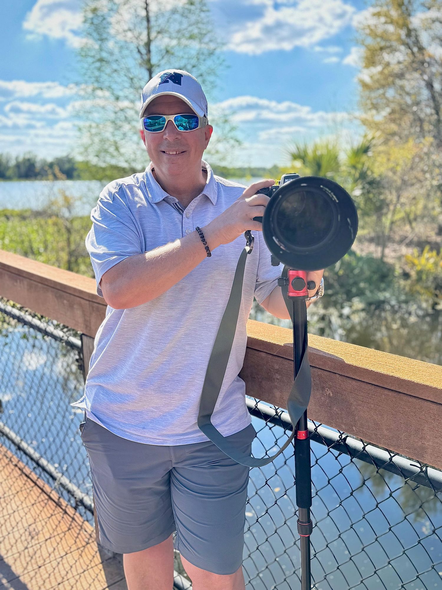 Photographer with camera gear stands on wooden deck overlooking lake on sunny day with blue skies and trees.