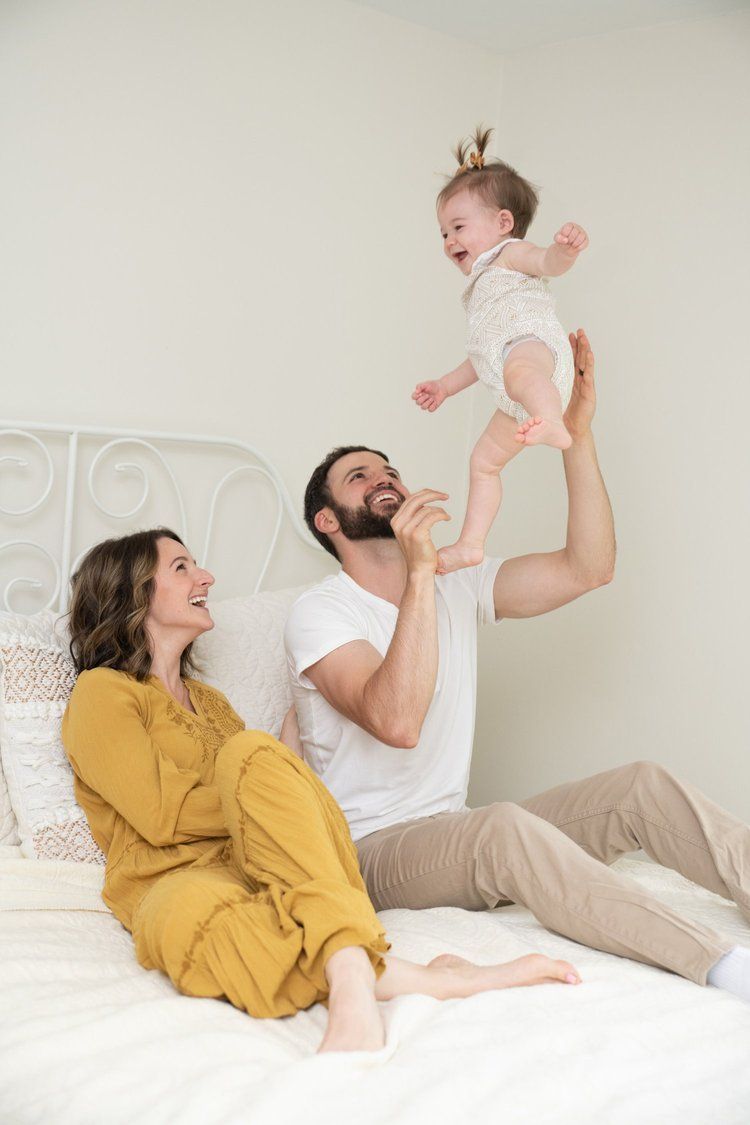 Family plays together on white bedding while wearing casual clothing and enjoying quality time together.