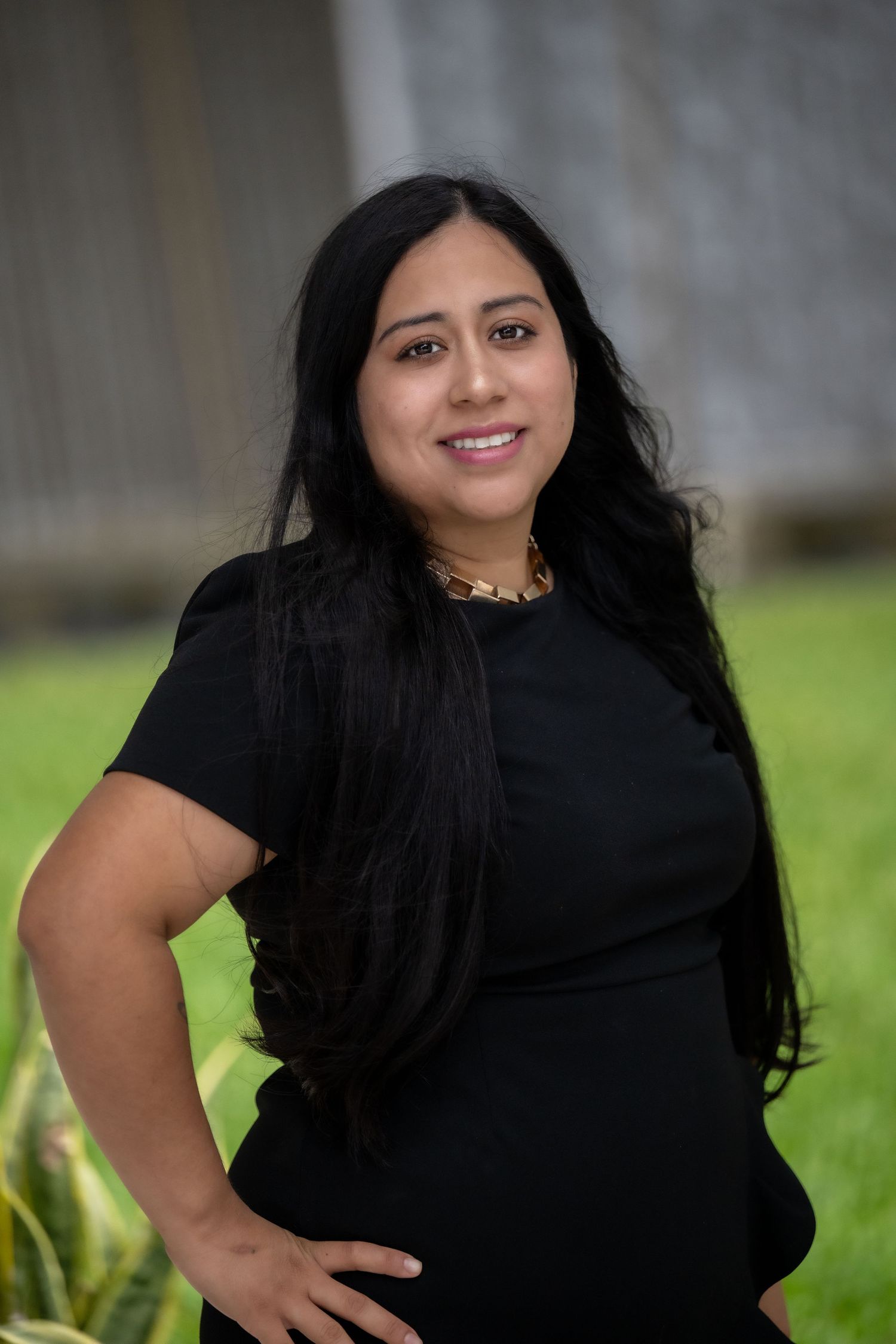 Smiling woman with long black hair wearing a black dress and gold necklace outdoors.