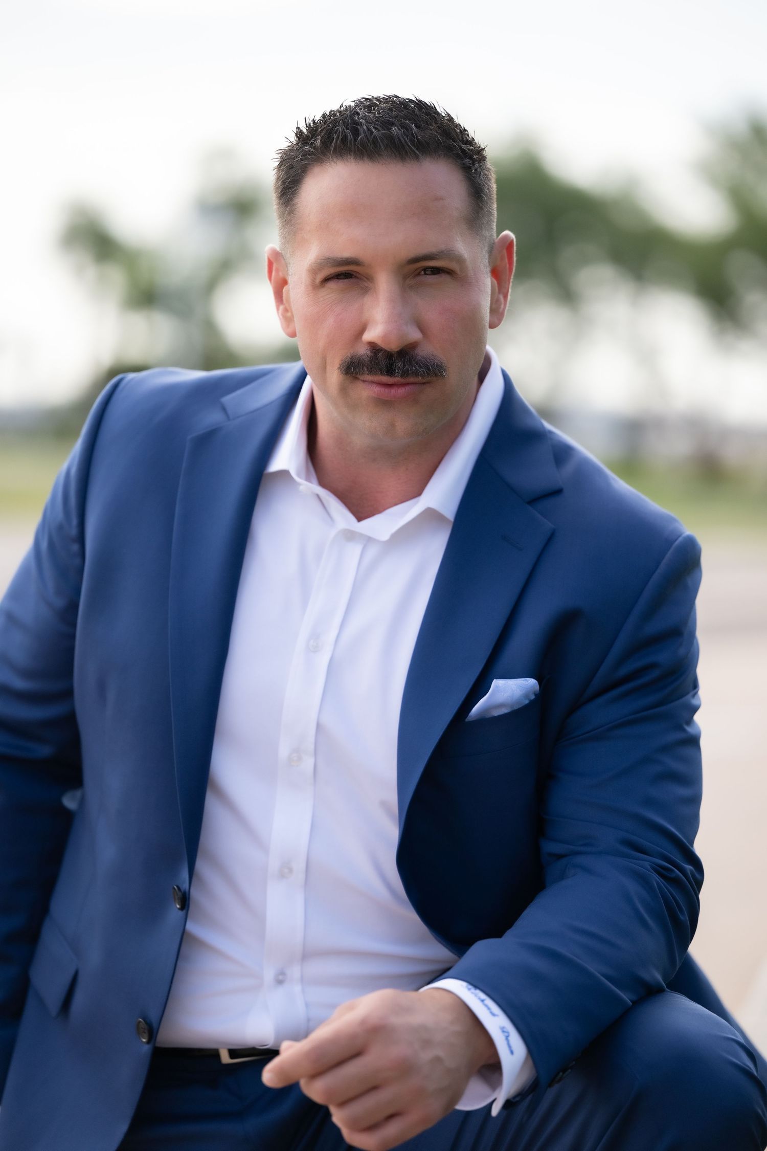 Professional man in blue suit with mustache poses outdoors in a confident business portrait photo.