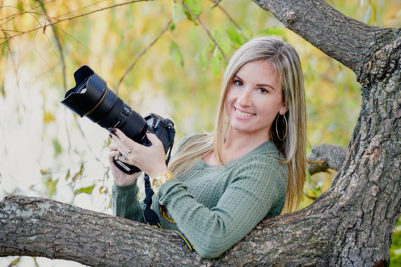 A person in a green sweater poses outdoors against autumn trees for a professional photo session.