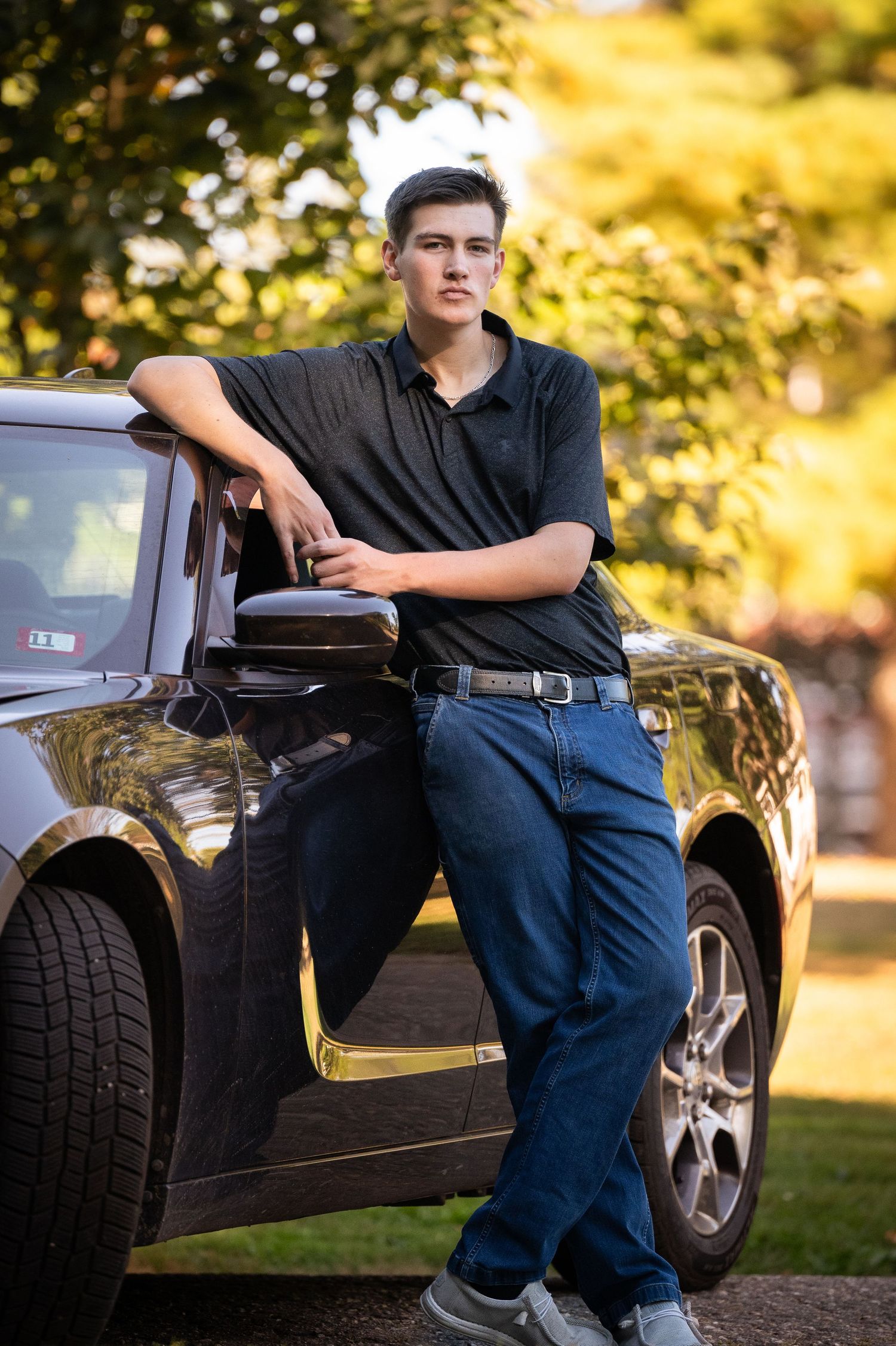 A person in dark clothing leans against a black car in a park setting during golden hour.