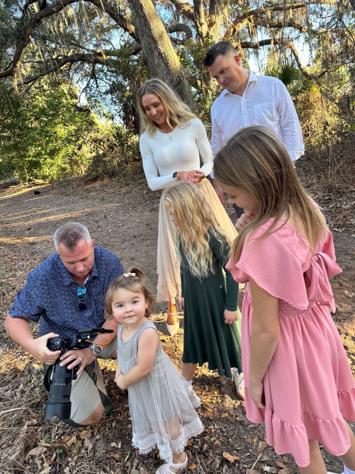 A group gathers under Spanish moss-draped trees on a dirt path while looking at something together on a sunny day.