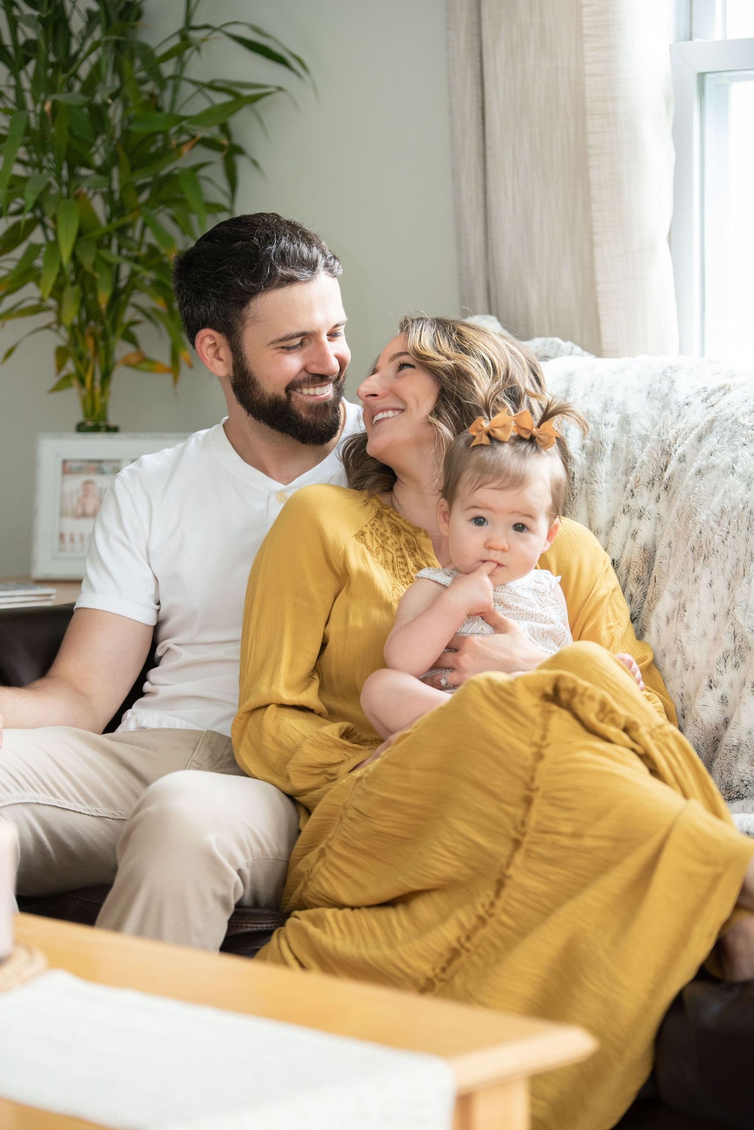 A family relaxes together on a couch wrapped in a yellow blanket in a bright living room with plants.