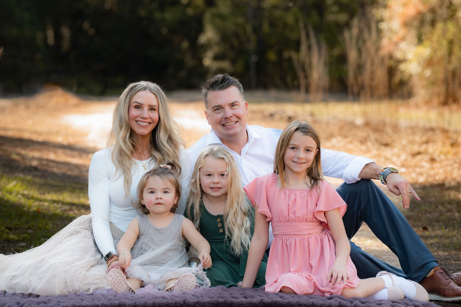 A family in coordinated casual outfits sits together on grass during an outdoor photo session at sunset.